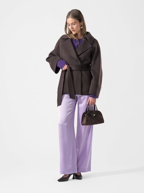 Una donna con un cappotto marrone e pantaloni viola tiene in mano una piccola borsa marrone e guarda in basso. Indossa scarpe nere e maniche viola spuntano da sotto il cappotto.