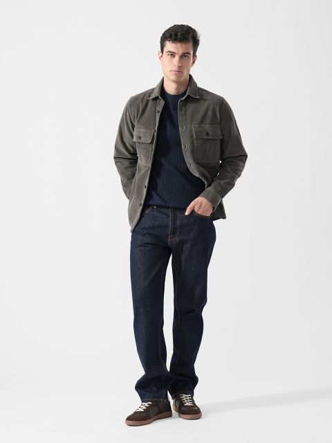 Un uomo con una giacca grigia, jeans blu scuro e sneakers marroni sta in piedi davanti a uno sfondo bianco semplice e ha le mani in tasca.