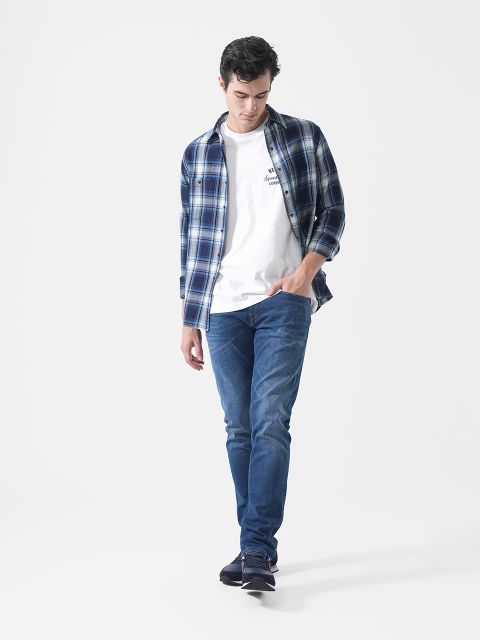 Un uomo con una camicia a quadri blu, una maglietta bianca, jeans blu e scarpe da ginnastica, che cammina davanti a uno sfondo bianco semplice e guarda in basso.