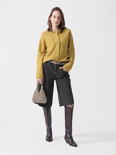 Una donna che indossa un maglione giallo, pantaloncini neri al ginocchio e stivali marroni, e che tiene in mano una borsa intrecciata, è in piedi davanti a uno sfondo bianco.