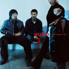 Due uomini sono seduti su una panchina, mentre una persona in primo piano tiene in mano una borsa porta abiti con la scritta "HUGO RED MEANS GO".