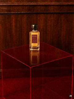 Su un tavolo rosso trasparente, davanti a una parete di legno e a un tappeto rosso, c'è una bottiglia di profumo Bel-Ami.
