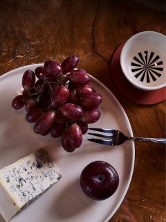 Un piatto di uva rossa, una fetta di formaggio erborinato e una prugna; inoltre, una forchetta e una tazza sono visibili su un tavolo di legno.