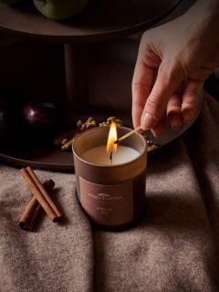 Una mano accende una candela marrone "African Tea" di Finch Hatton, che si trova accanto a bastoncini di cannella, prugne e noci su un morbido panno marrone.