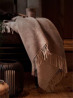 Una coperta beige con frange drappeggiata su una sedia, accanto una candela in una lanterna nera e una tazza di caffè sul pavimento di legno.