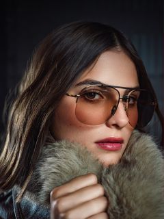 Una donna con lunghi capelli castani indossa occhiali da aviatore colorati e un cappotto con collo di pelliccia, guardando con sicurezza la telecamera su uno sfondo scuro.