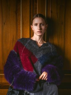 Una donna con un maglione soffice a strisce rosse, blu e viola è in piedi davanti a una parete di legno, tiene in mano una borsa e guarda la fotocamera.