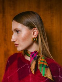 Donna con un maglione rosso, una sciarpa a quadri colorata e orecchini d'oro, che guarda a sinistra, su uno sfondo di legno.