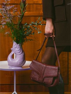 Una persona in abiti scuri tiene una borsa bordeaux accanto a un vaso viola a forma di pesce con fiori su un tavolo bianco.