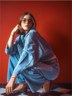 Una donna con occhiali da sole e giacca di jeans è accovacciata su un pavimento a scacchi bianchi e neri davanti a un muro rosso.