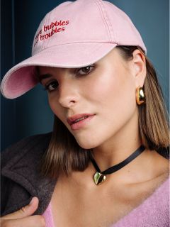 Una donna con capelli lisci e castani indossa un cappello rosa, orecchini d'oro, una collana a cuore e un top rosa e soffice, e guarda la telecamera davanti a uno sfondo blu.