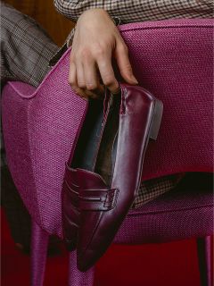 Una persona in pantaloni a quadri è seduta su una sedia rosa con una superficie strutturata e tiene in mano una pantofola bordeaux.
