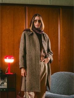 Una persona con un cappotto marrone e una sciarpa si trova in una stanza accanto a una lampada rossa e una sedia grigia, con pareti rivestite in legno sullo sfondo.