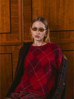 Una donna che indossa un maglione rosso a rombi, occhiali da sole e orecchini d'oro è seduta davanti a una parete con pannelli in legno e guarda la telecamera.