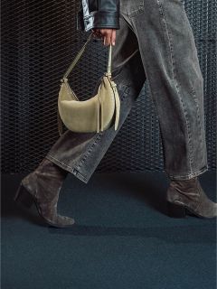 Una persona in jeans grigi e stivali scamosciati con tacco tiene una borsa beige e cammina davanti a uno sfondo scuro strutturato.