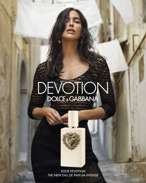 Una donna vestita di nero si trova in uno stretto vicolo dietro una bottiglia di profumo "Dolce & Gabbana Devotion". Sullo sfondo è appesa della biancheria bianca.