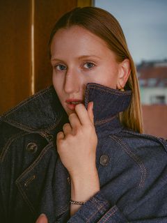 Una donna con i capelli lisci indossa una giacca di jeans scura con il colletto alzato e guarda la telecamera con un'espressione impassibile.
