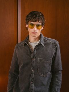 Un uomo con i capelli mossi, che indossa occhiali da sole gialli, una camicia di jeans grigia e un maglione chiaro, è in piedi davanti a una parete di legno e guarda la fotocamera.