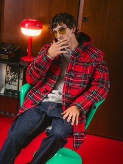 Un uomo con un cappotto a quadri rosso e occhiali da sole è seduto su una sedia verde accanto a una lampada rossa e tiene la mano vicino al viso in una posa elegante.