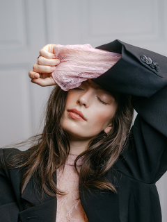 Una persona con un blazer nero e un top di pizzo rosa posa tenendosi una mano sulla fronte.