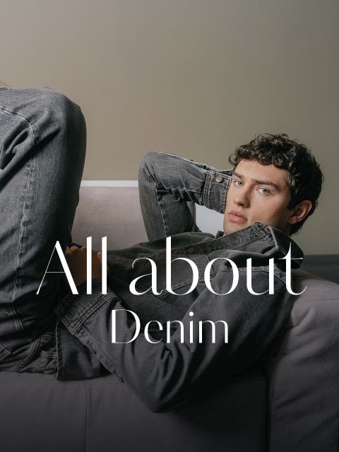 Un giovane uomo con una giacca di jeans grigia e jeans è appoggiato a un divano, sull'immagine c'è la scritta "All about Denim".
