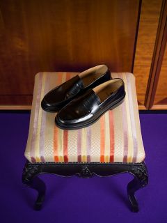 Un paio di mocassini in pelle nera sono appoggiati su uno sgabello imbottito a righe con gambe nere decorate, che poggia su un tappeto viola.
