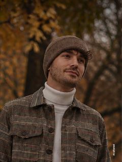 Un uomo con un cappello marrone e una giacca a quadri è in piedi all'aperto sullo sfondo di alberi autunnali, guarda di lato e sorride leggermente.