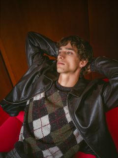 Una persona con i capelli ricci, che indossa una giacca di pelle e un maglione argyle, si rilassa su una poltrona rossa, appoggiandosi all'indietro e con le mani incrociate dietro la testa.