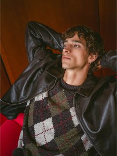 Su uno sfondo di legno, un uomo con una giacca di pelle e un maglione argyle si appoggia all'indietro con le mani dietro la testa, guardando in alto.