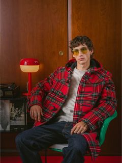 Un uomo con un cappotto a quadri rosso e occhiali da sole gialli è seduto su una sedia verde, con una lampada rossa e una parete di legno sullo sfondo.