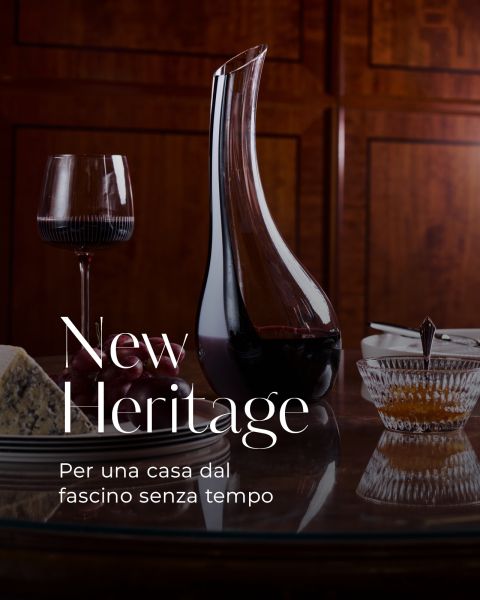 Su un tavolo lucido con pannelli in legno ci sono una caraffa, un bicchiere da vino, del formaggio e una ciotola; il testo recita: "New Heritage. Per una casa dal fascino senza tempo.".