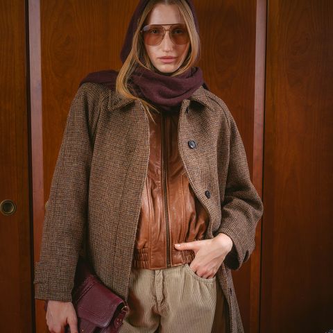 Una donna con occhiali da sole, una sciarpa marrone, un cappotto e una giacca di pelle, che tiene una borsa e sta in piedi davanti a uno sfondo di legno.