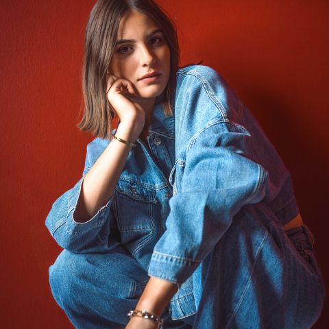 Una donna in giacca e jeans di denim è seduta davanti a un muro rosso brillante, appoggiando il mento sulla mano e guardando la fotocamera.