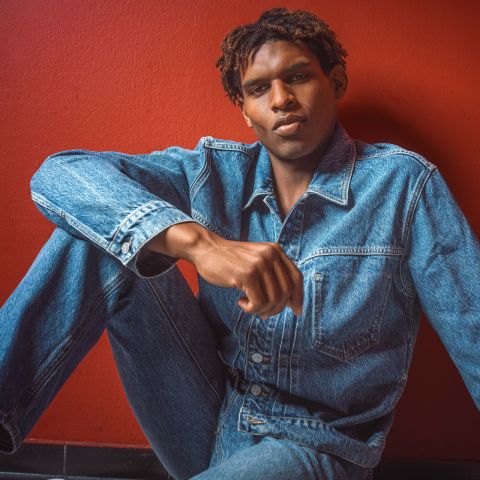 Una persona con giacca e jeans di denim è seduta davanti a una parete rosso vivo e guarda la fotocamera con un'espressione impassibile.