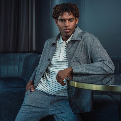 Un uomo in abito grigio e camicia a righe è seduto su un divano blu, appoggiando un braccio su un tavolo di vetro e guardando con sicurezza la telecamera.