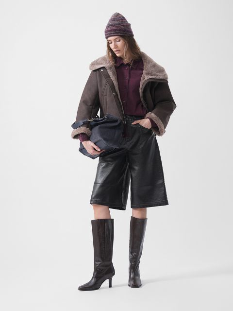 Una donna con cappello, giacca di montone, camicia bordeaux, culotte di pelle e stivali con tacco, che tiene in mano una borsa blu navy e si trova davanti a uno sfondo bianco.
