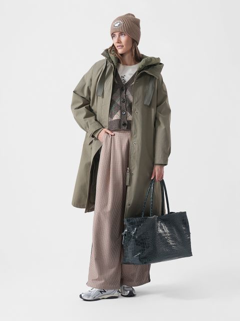 Una donna con un cappotto verde, pantaloni fantasia, sneakers e un cappello tiene una grande borsa strutturata e si trova di fronte a uno sfondo bianco semplice.