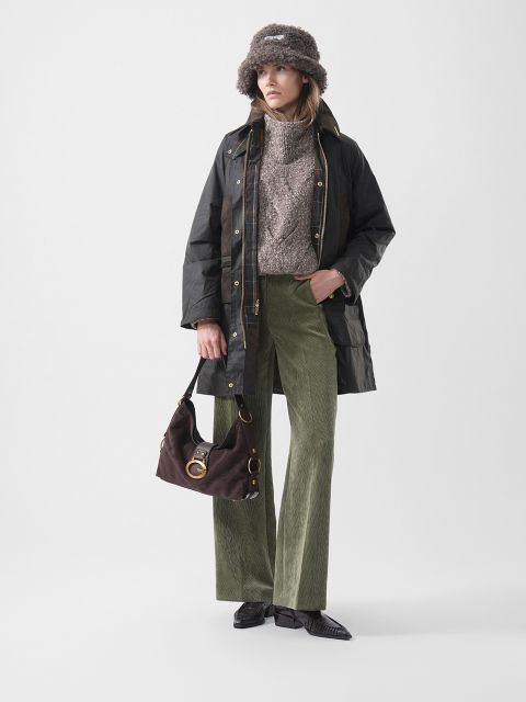 Una donna con un cappello di pelliccia, una giacca scura, un maglione marrone, pantaloni di velluto a coste verdi e scarpe nere, che tiene una borsa marrone e sta in piedi davanti a uno sfondo semplice.