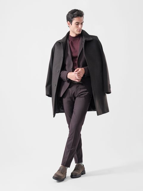 Un uomo che indossa un cappotto scuro sulle spalle, un abito bordeaux, un maglione bordeaux e scarpe marroni, in piedi davanti a uno sfondo semplice e chiaro.
