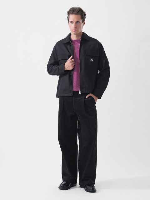 Un uomo posa con una giacca nera sopra un maglione viola, pantaloni neri a gamba larga e scarpe nere, su uno sfondo bianco semplice.