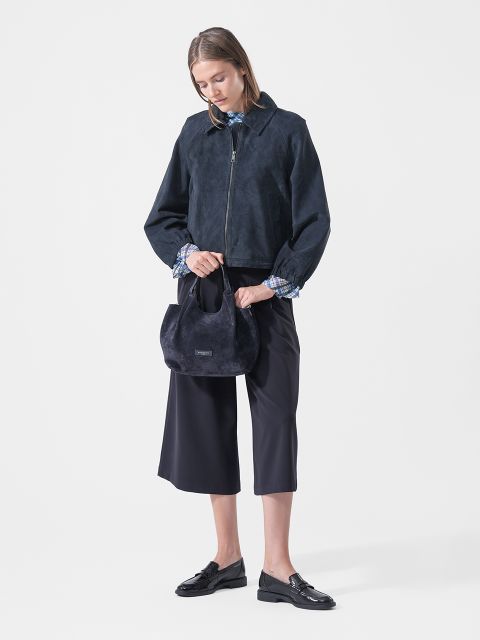 Una donna con una giacca blu navy, culotte e mocassini tiene una piccola borsa blu navy e si trova di fronte a un semplice sfondo bianco.