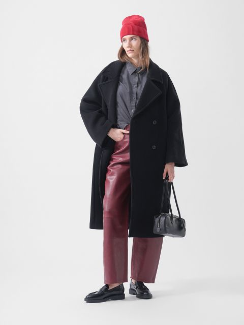 Una donna con un berretto rosso, un cappotto nero, una maglietta grigia, pantaloni di pelle rossi e mocassini neri tiene una borsa nera e posa davanti a uno sfondo bianco.