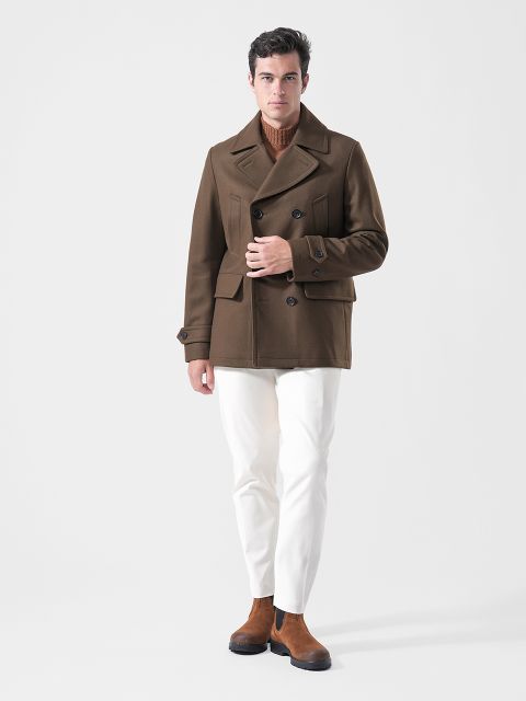 Un uomo che indossa un cappotto marrone, un maglione marrone chiaro, pantaloni bianchi e stivali marroni è in piedi davanti a uno sfondo bianco semplice.