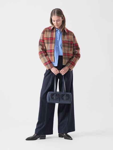 Una donna con una giacca a quadri, una camicia blu e pantaloni a righe sottili larghe tiene in mano una borsa blu navy a due scomparti e si trova di fronte a uno sfondo bianco semplice.