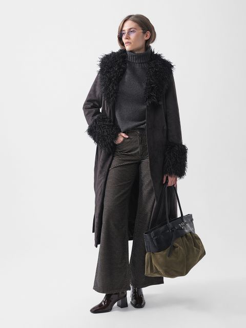 Una donna è in piedi, vestita con un cappotto scuro bordato di pelliccia, un dolcevita, pantaloni a zampa d'elefante e stivali. Indossa una borsa bicolore e occhiali, e posa con sicurezza.