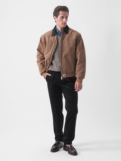 Un uomo che indossa una giacca marrone, un maglione grigio, una camicia blu, pantaloni neri e scarpe scure, in piedi davanti a uno sfondo bianco semplice.