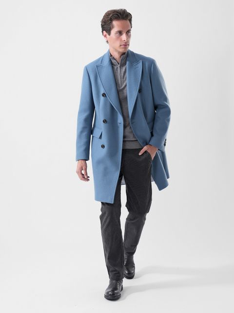 Un uomo in un abito doppiopetto azzurro, una camicia grigia, pantaloni scuri e stivali neri, che cammina con sicurezza davanti a uno sfondo bianco semplice.