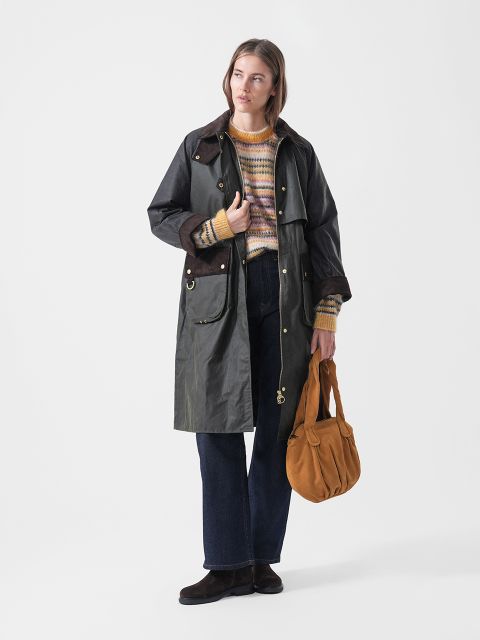 Una donna con un cappotto scuro, un maglione a righe, jeans blu e scarpe marroni, che tiene in mano una borsa color senape e sta in piedi davanti a uno sfondo bianco semplice.