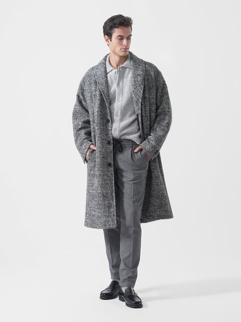 Un uomo che indossa un cappotto grigio testurizzato, una camicia abbottonata grigio chiaro, pantaloni grigi e scarpe nere è in piedi con le mani in tasca davanti a uno sfondo bianco.