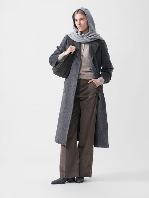Una donna con un cappotto grigio con cappuccio, un maglione beige, pantaloni marroni a gamba larga e scarpe nere, che tiene una borsa nera e sta in piedi su uno sfondo bianco.